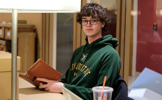 Male student sitting at table with lamp reading book in archives