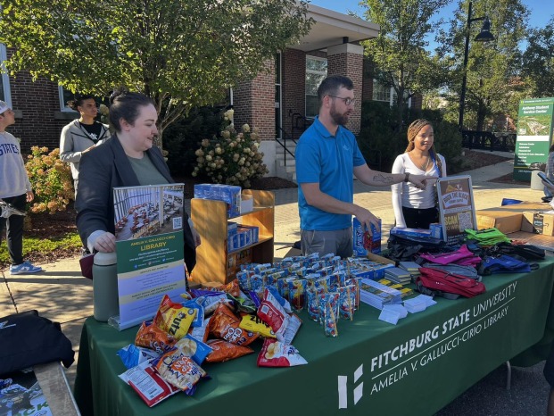 Library staff standing behind a table covered with library promotional materials, snacks, and stationery at an outdoor event at Fitchburg State University. The tablecloth displays the university's library name.